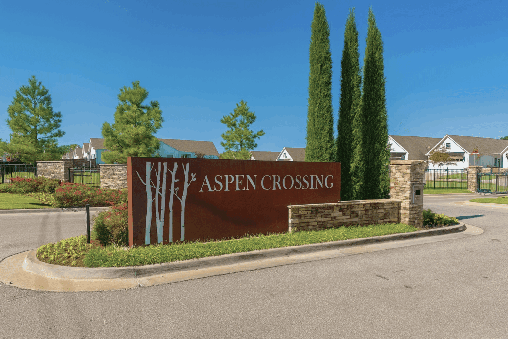 Monument entrance sign for Aspen Crossing Patio Homes in Broken Arrow, OK, with stone columns, landscaping, and gated access in the background.