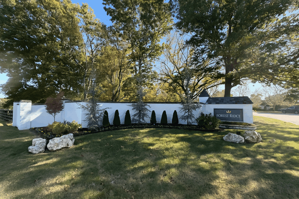 Entrance monument at Forest Ridge neighborhood in Broken Arrow, Oklahoma, with manicured landscaping, evergreen trees, and white brick wall under sunny skies.