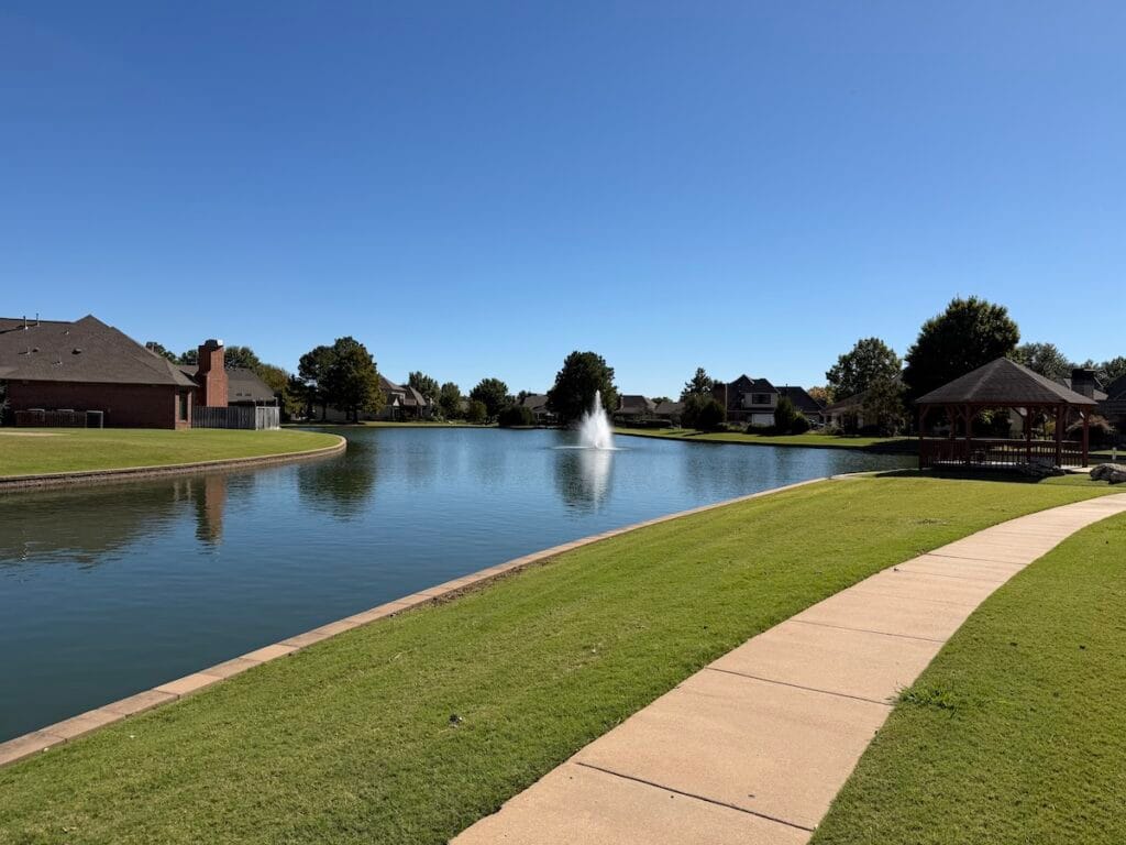 A peaceful lakeside walking path overlooking a fountain and gazebo in The Lakes at Indian Springs neighborhood in Indian Springs Broken Arrow.
