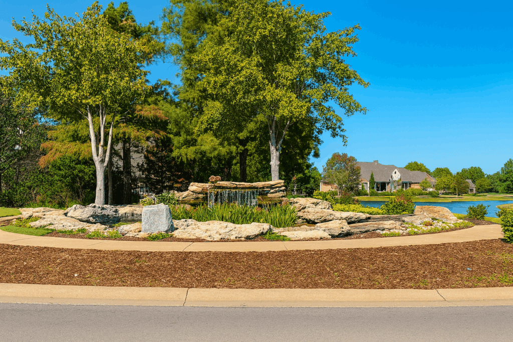 A landscaped stone waterfall surrounded by trees and well-kept lawns inside The Lakes at Indian Springs, a neighborhood in Indian Springs Broken Arrow.